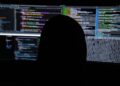 Silhouette of a person working in front of computer monitors displaying code, used as an illustrative image for cross-border cybercrime reporting.