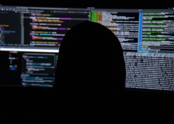 Silhouette of a person working in front of computer monitors displaying code, used as an illustrative image for cross-border cybercrime reporting.