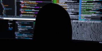 Silhouette of a person working in front of computer monitors displaying code, used as an illustrative image for cross-border cybercrime reporting.
